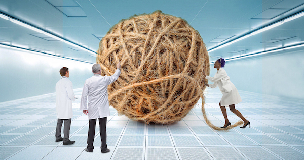 Giant ball of brown twine sits in a clean room, which is all white and very sterile looking. Three scientists in lab coats look on.