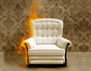 Photo of arm chair in flames. A white upholstered arm chair, which is on fire, sits against a wall covered in classic floral-like wall paper.