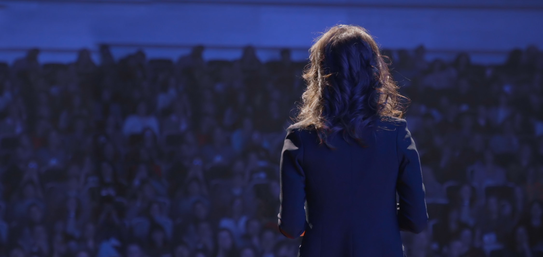 Woman standing on a stage at a national sales meeting with the spotlight on her, her back is to the camera, she faces a full audience in a dark auditorium