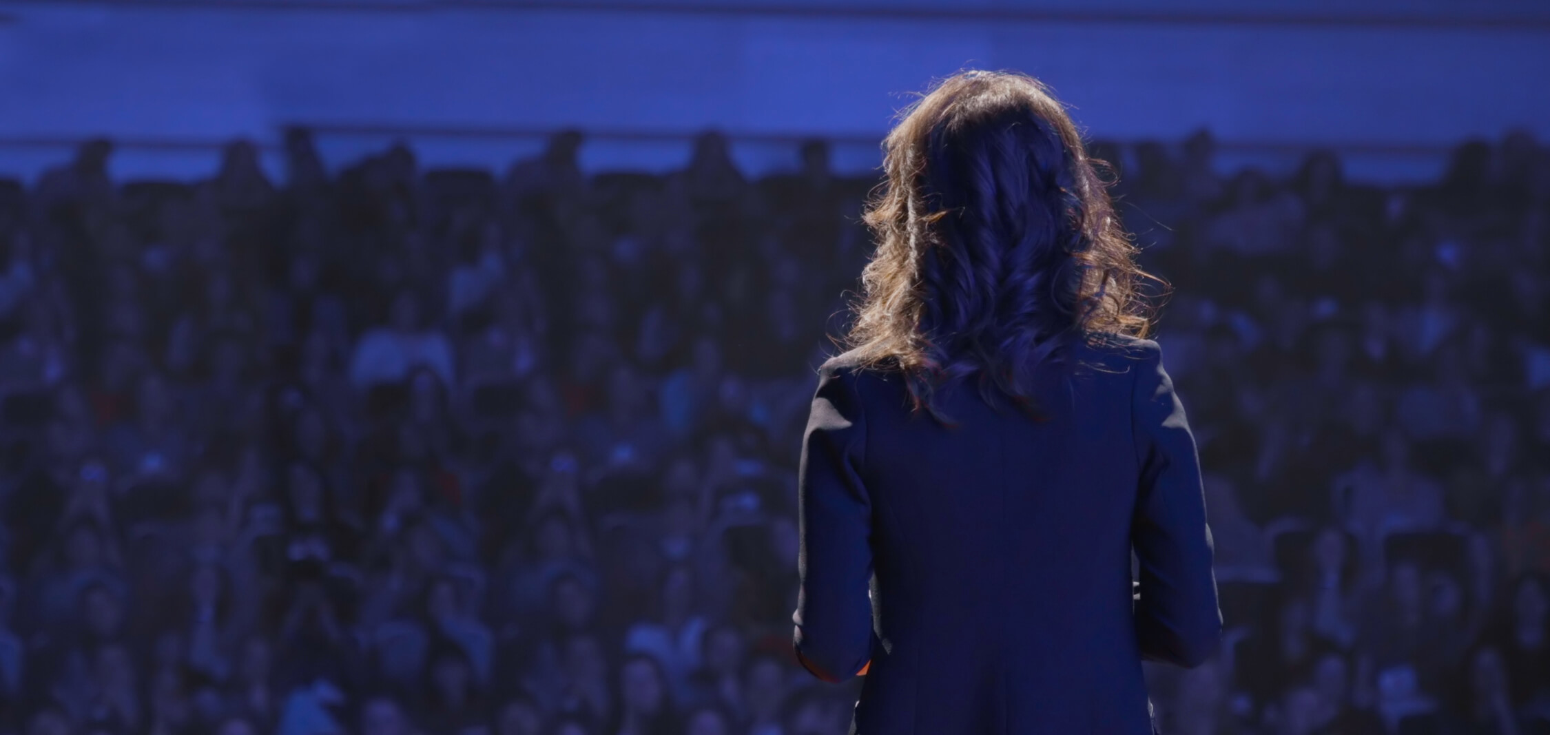 Woman stands with her back to the reader in a dark space as if she's in front of a crowd on stage. A light shines on her.