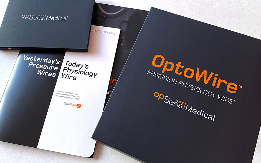 Photo of sales collateral displayed in a fan across a table. Up top left is a small black folder that says OpSense Medical. Below is the blakc and white brochure that says, "Yesterday's Pressure Wires. Today's Physiology Wire. (additional text that is illegible)" On left is a square black box that says "OptoWire™ Precision Physiology Wire™ OpSens Medical